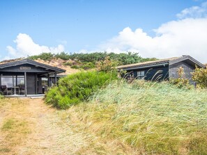 A building with a dark exterior, a covered outdoor seating area, and a natural landscape with grass and shrubs. - 6 Person Holiday Home in Henne (Henne)