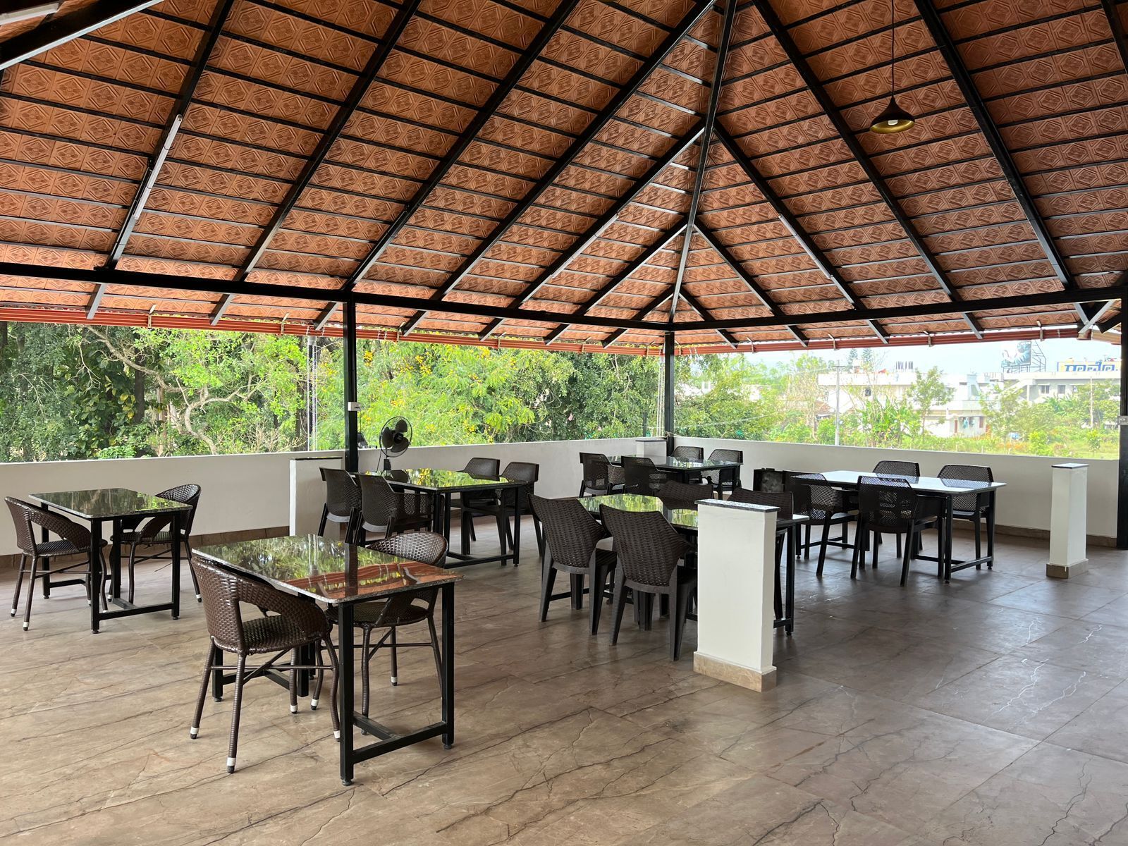 Un patio al aire libre con mesas y sillas, techo de paja y vistas a zonas verdes y edificios.