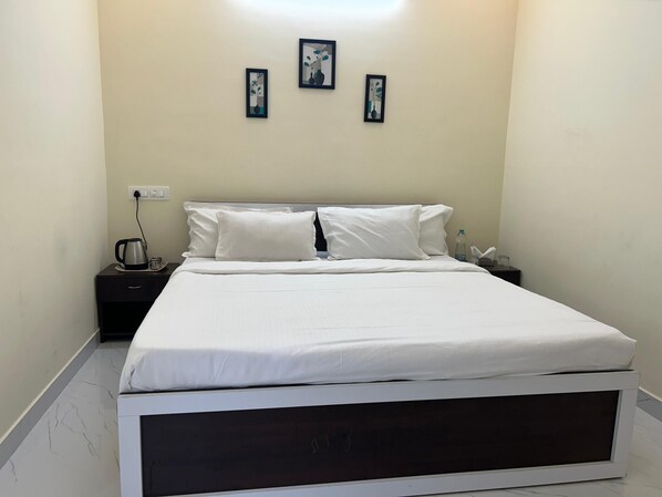 A neatly made bed with white linens, a dark wooden headboard, and a nightstand with a kettle and a bottle of water. Three framed artworks are hung on the wall above the bed. - Sriram Guest House (Chikkamagaluru)