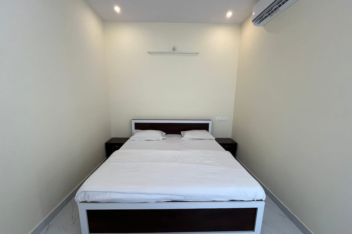 Een eenpersoonsbed met een witte matras en donker hoofdbord in een kleine, smalle kamer met witte wanden en een witte vloer.