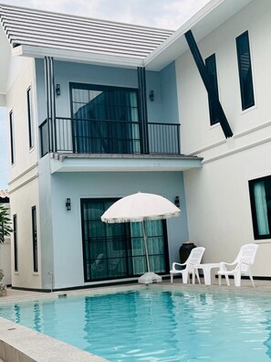 A modern two-story building with a swimming pool, a balcony with a white umbrella, and white outdoor furniture. - Level FIVE pool villa Pattaya (Bang Lamung)