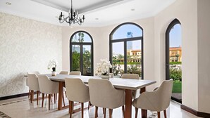 A dining area with a large table, six chairs, and a chandelier. There are three large windows with arched tops, and a view of buildings and greenery outside.