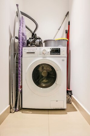 A front-loading washing machine with a control panel, a hose, and a mop leaning against the wall. - FIRST CLASS | 1BR | Stylish Gem | Near Dubai Mall (Downtown)