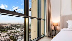 A hotel room with a large window offering a city view, a bed with white linens, and a bedside table with a lamp.