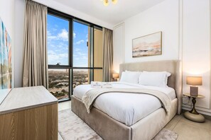 A hotel room with a large bed, a desk, and a view of the city through large windows.