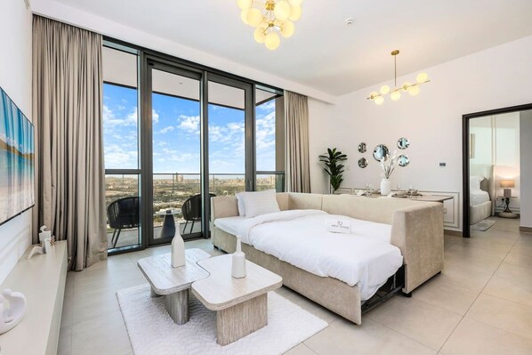 A modern hotel room with a large window, a sofa, a dining table, and a TV.