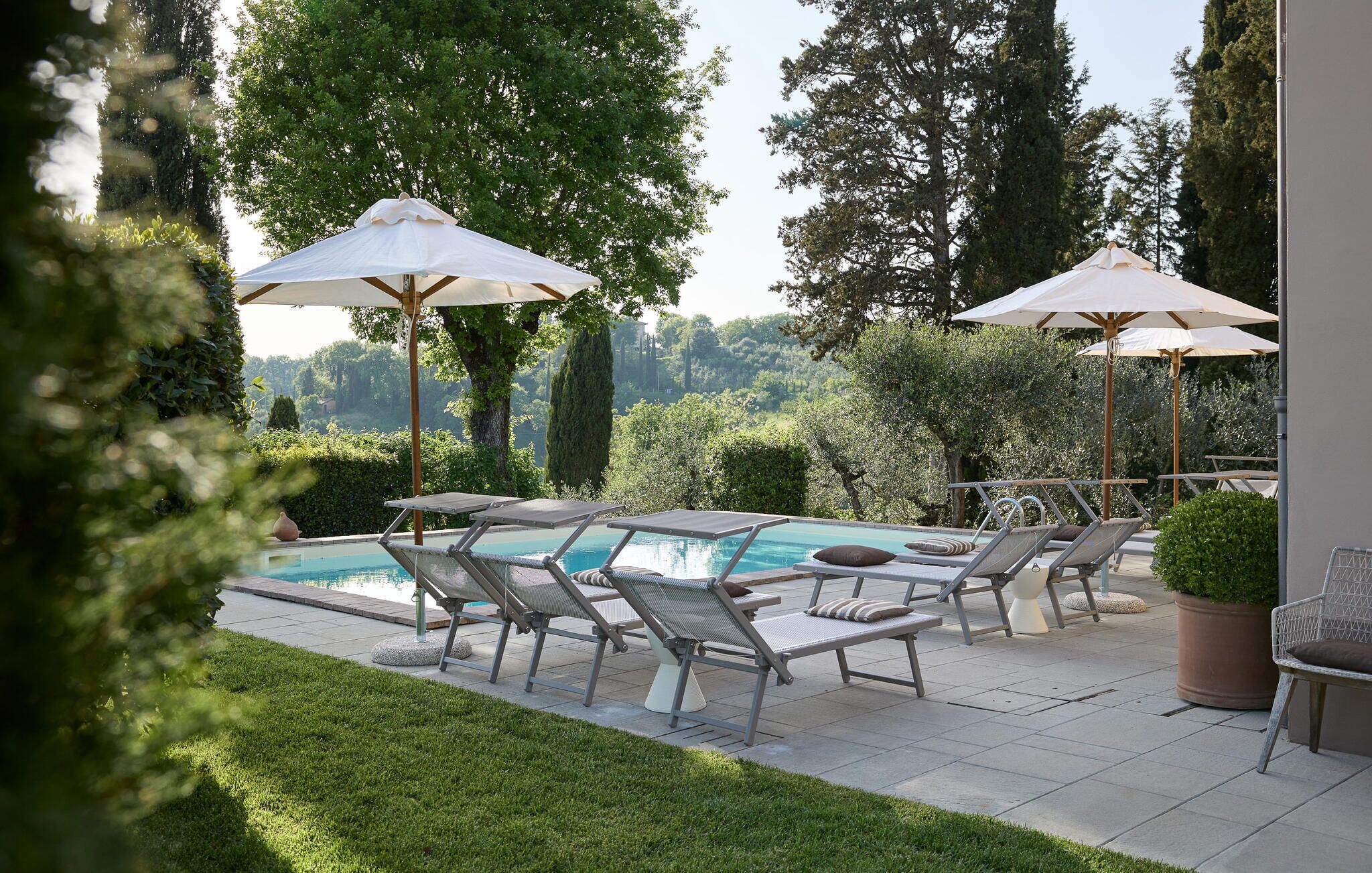 Zona junto a la piscina con sillones de descanso y sombrillas, rodeada de follaje y árboles.