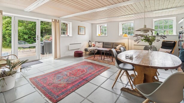 A spacious living area with a round wooden table, a sofa, and a dining table. There are multiple windows providing natural light and a view of greenery outside. - Scenic area close to forest, golf, and beach (Rønne)
