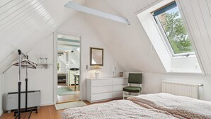 A bedroom with a sloped ceiling, a bed with floral bedding, a dresser, a mirror, and a window with a view of trees. - Scenic area close to forest, golf, and beach (Rønne)