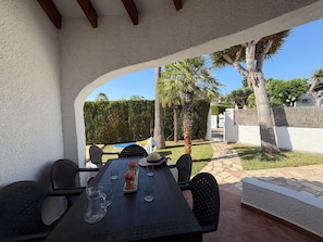 An outdoor patio with a table set for dining, surrounded by chairs, and a view of a garden with palm trees and a hedge.