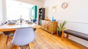 Un coin repas avec une table en bois, des chaises, un banc, une plante en pot, une bibliothèque et une horloge au mur.