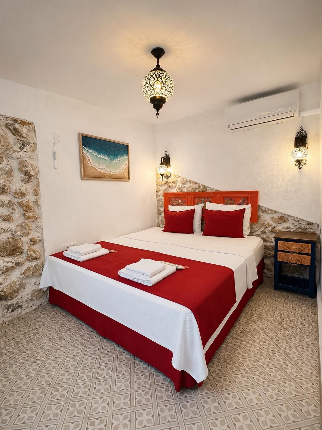 Un dormitorio con pared de piedra, cama con ropa de cama roja y blanca, un cuadro de playa y suelo estampado.