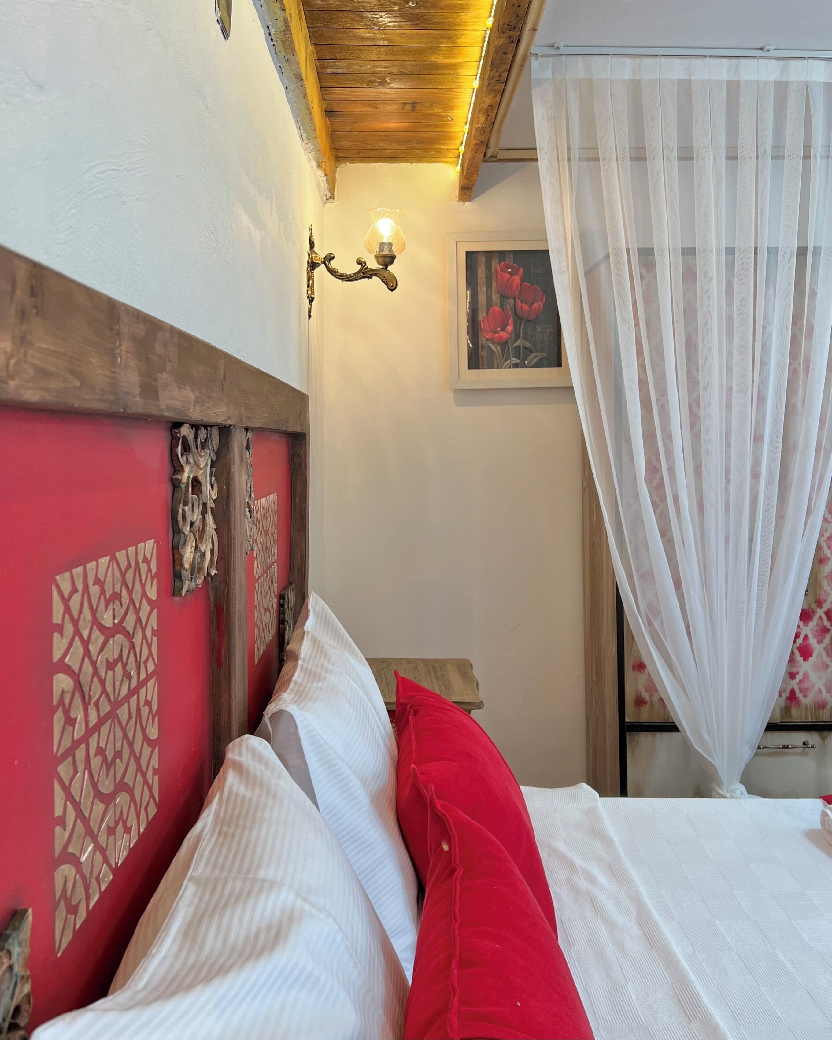 Un dormitorio con cabecera de madera, una mesita de noche con lámpara, ropa de cama blanca y roja, y una ventana con cortina translúcida.