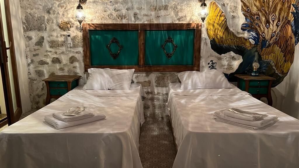 Ein Zimmer mit zwei Betten, einer Steinwand, einem türkisblauen Kopfteil und einem Pfauen-Wandbild.