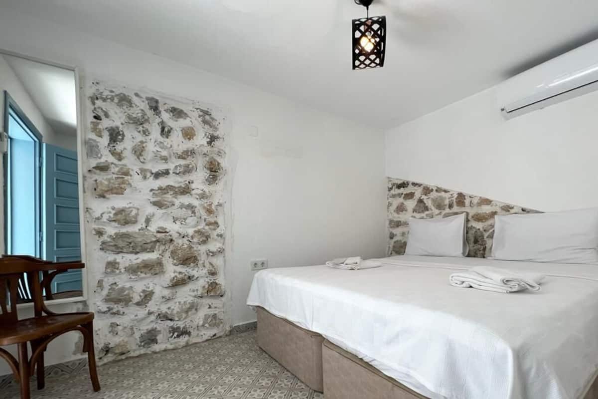 A bedroom with a stone wall, a bed with white linens, a wooden chair, and a patterned floor.