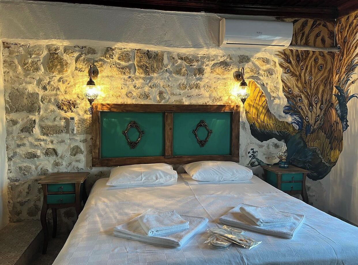 Una habitación con pared de piedra, una cama con ropa de cama blanca, dos mesitas de noche y un mural de pavo real.