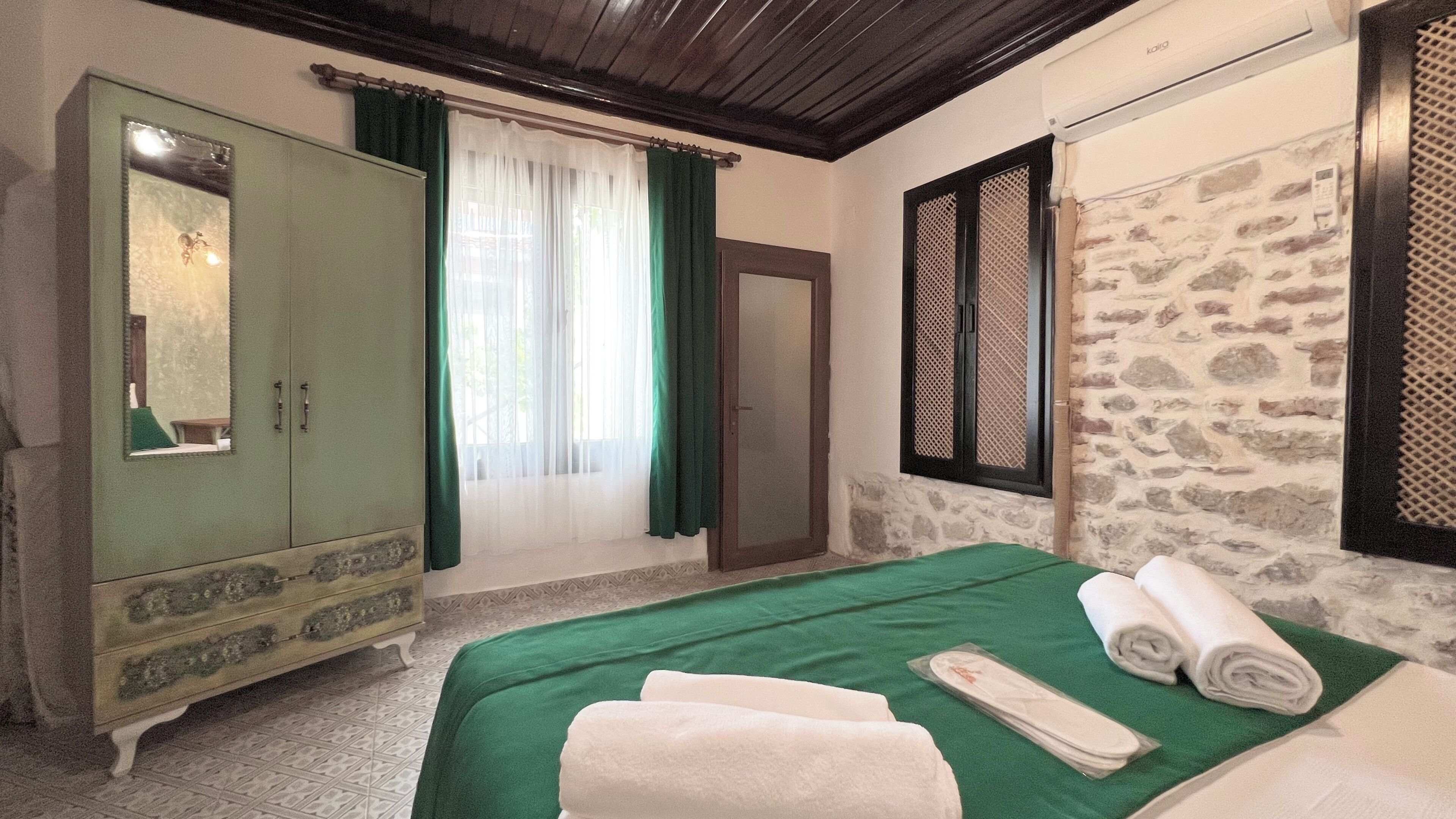 Una habitación con una cama, colcha verde, armario de madera, espejo, ventana con cortinas y pared de piedra.
