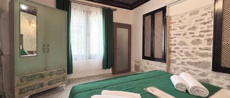 Una habitación con una cama, colcha verde, armario de madera, espejo, ventana con cortinas y pared de piedra.