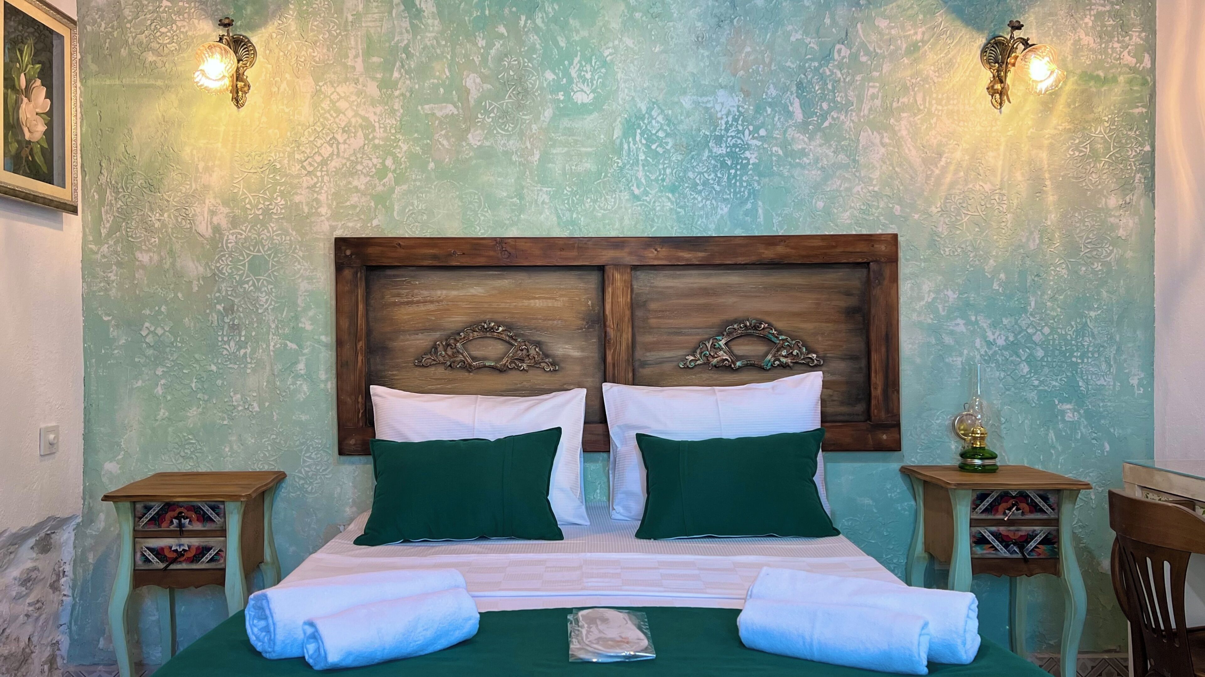 Uma cama de casal com roupa de cama verde e branca, duas mesas de cabeceira com objetos decorativos e uma parede com pintura texturizada e arandelas.