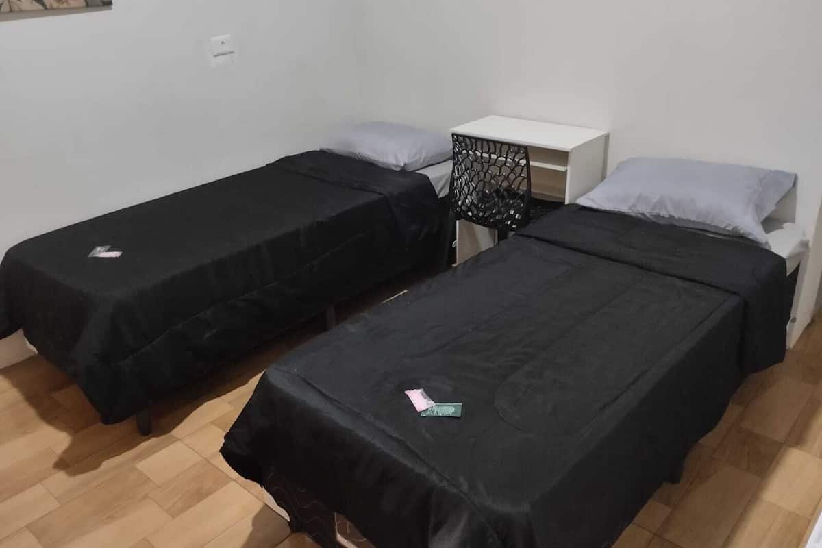 A room with two single beds, a small white nightstand, a black chair, and a wall-mounted shelf.