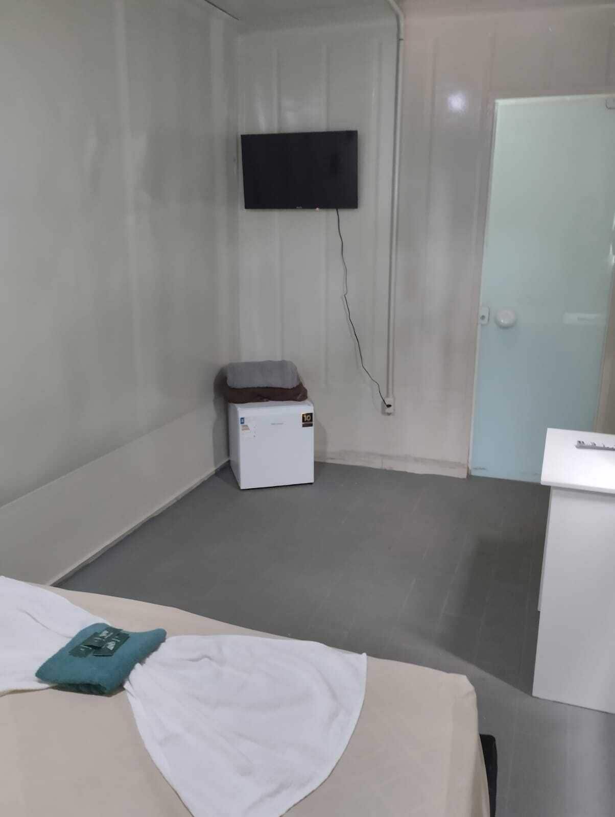 A small, clean room with a bed, a small white cabinet, a television mounted on the wall, and a door.