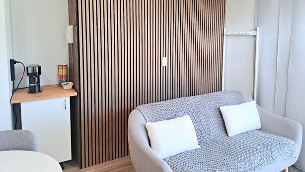 Een moderne kamer met een grijze bank, een houten lattenwand, een klein houten tafeltje met een koffiezetapparaat en een witte stoel.