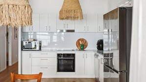 A modern kitchen with white cabinets, stainless steel appliances, and a wooden dining table. - Sunset Cottage on the Burrum River (Burrum Heads)