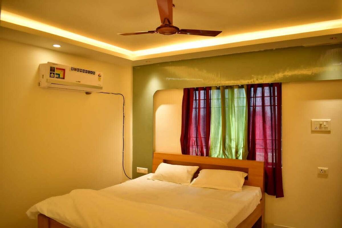 Habitació d'hotel amb llit, ventilador de sostre, aire condicionat i finestra amb cortines de colors.