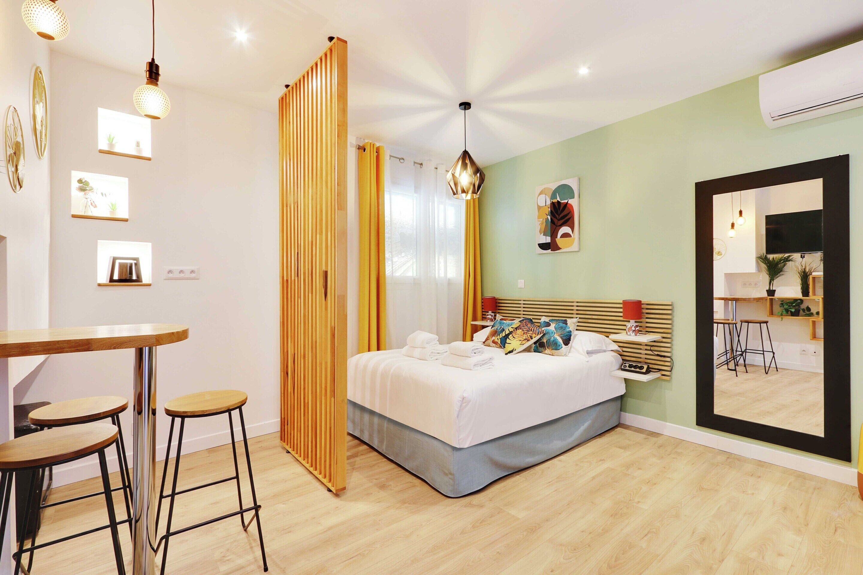 Une chambre moderne avec un lit, une cloison en bois, un coin repas avec des tabourets et un grand miroir.