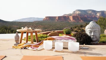 A serene outdoor setting with a table set for a tea ceremony, featuring a Buddha statue, a wooden table, and a scenic mountain backdrop.