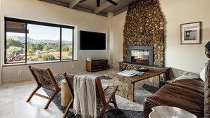A spacious living room with a stone fireplace, large windows offering a view of the outdoors, and a ceiling fan. - Sedona Infinity Pool Paradise | Red Rock Views | Hot Tub (Sedona)