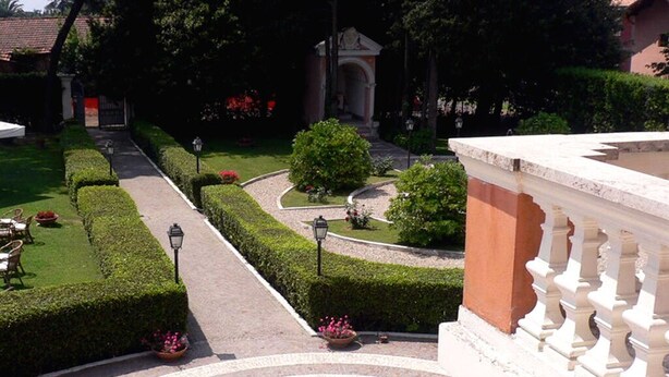 Een verzorgde tuin met een geplaveid pad, strak bijgesnoeide hagen en een klassiek gebouw op de achtergrond.