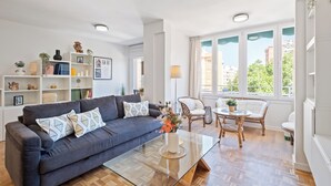 A modern living room with a blue sofa, glass coffee table, wicker chairs, and a bookshelf.