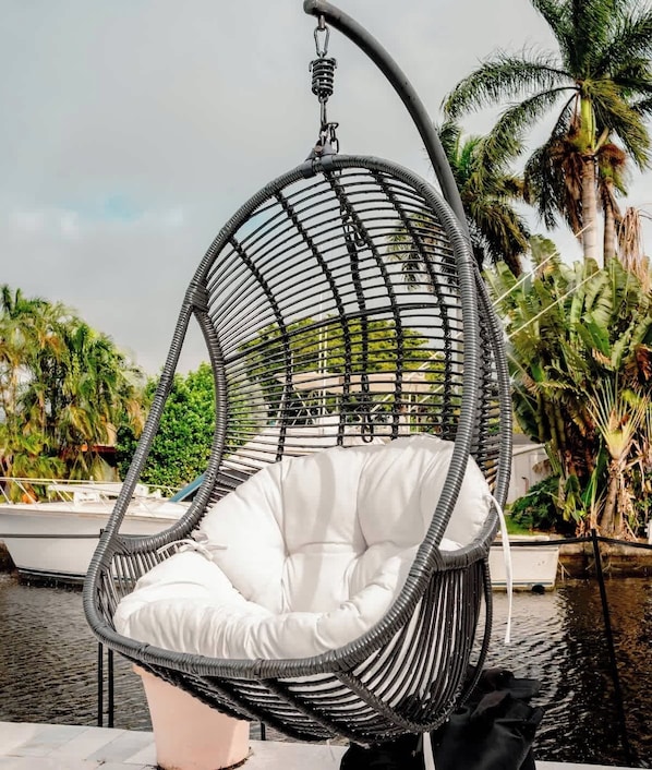 A black wicker hanging chair with a white cushion, a boat in the background, and palm trees.