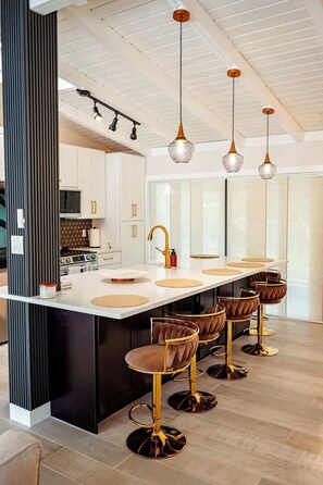 A modern kitchen with a central island, bar stools, pendant lighting, and a large window.