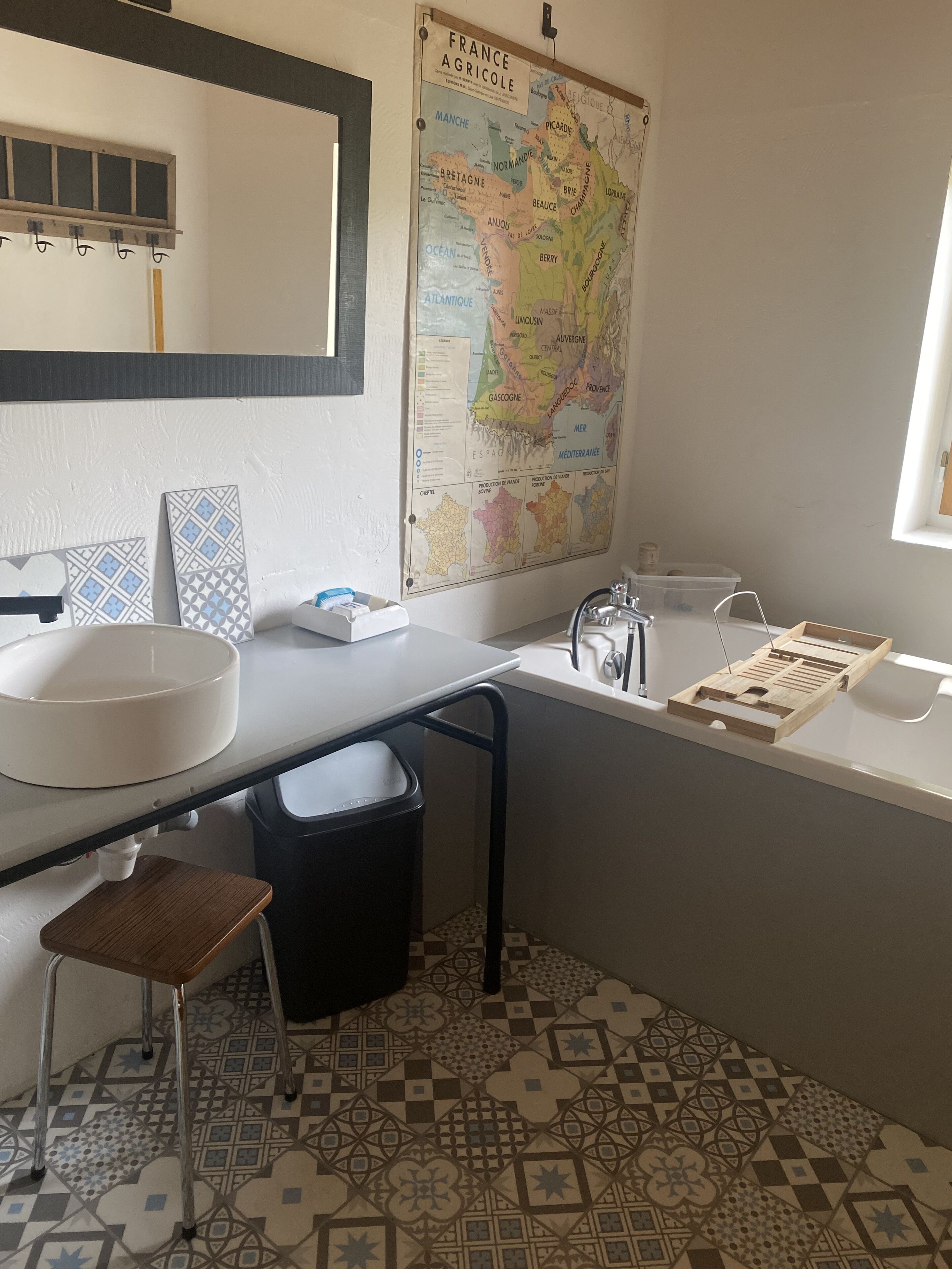 A bathroom with a large map of France, a white sink, a black trash bin, a wooden stool, and a bathtub.