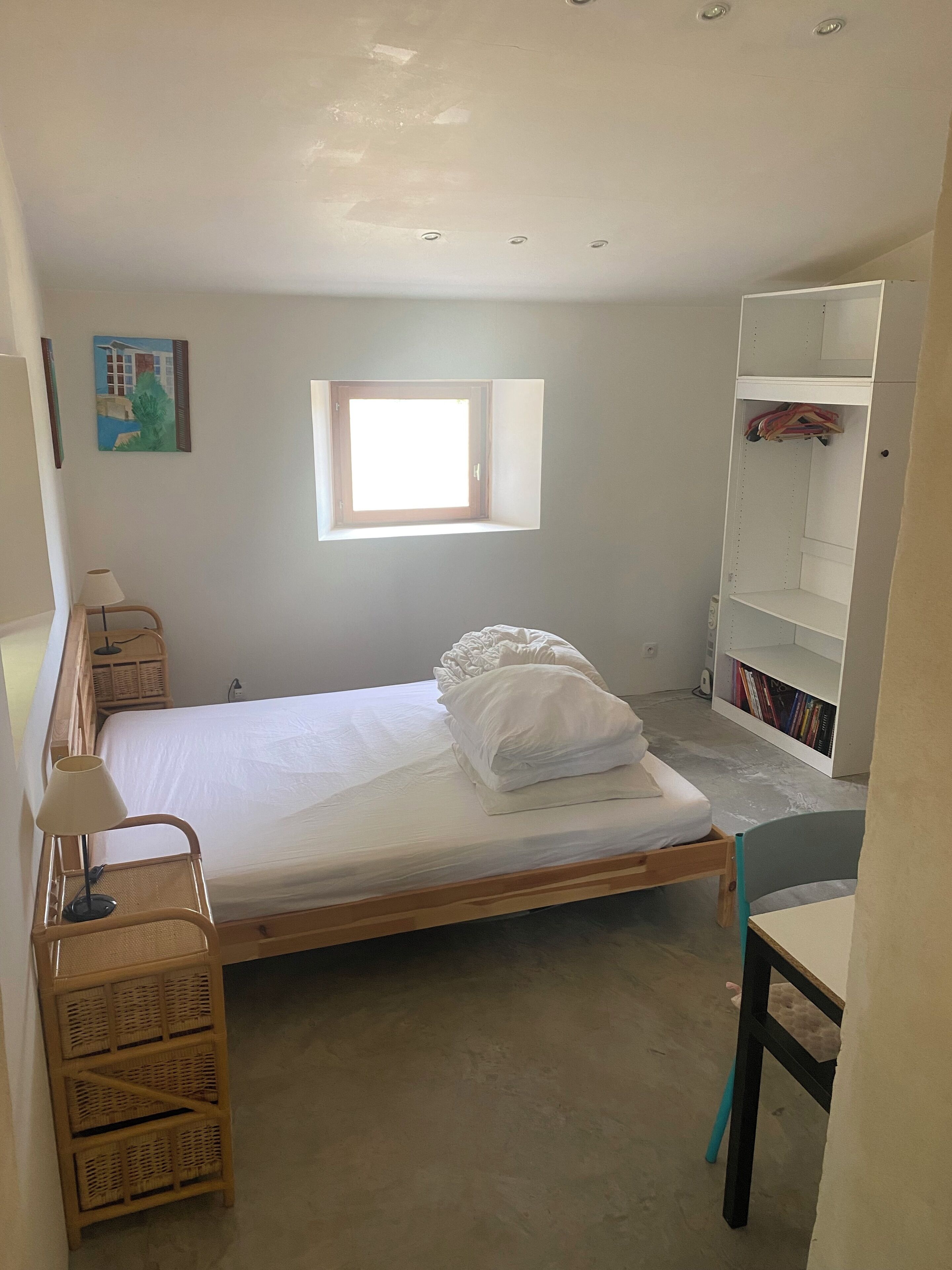 A bedroom with a single bed, a small window, a bookshelf, and a chair.