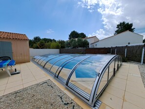 Un espace extérieur abrité, avec une table et des chaises, un petit abri de jardin et, au fond, un bâtiment blanc.