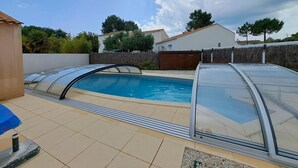 A swimming pool with a glass enclosure, surrounded by a tiled patio and a wooden fence. - Charming villa 6 pers, quiet residence, private pool (Saint-Jean-de-Monts)