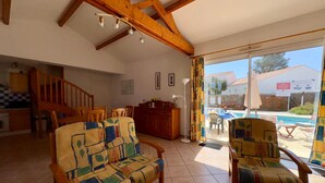 A spacious living area with a high ceiling, wooden beams, a sofa, chairs, a dining table, and a view of a swimming pool through a glass door. - Charming villa 6 pers, quiet residence, private pool (Saint-Jean-de-Monts)