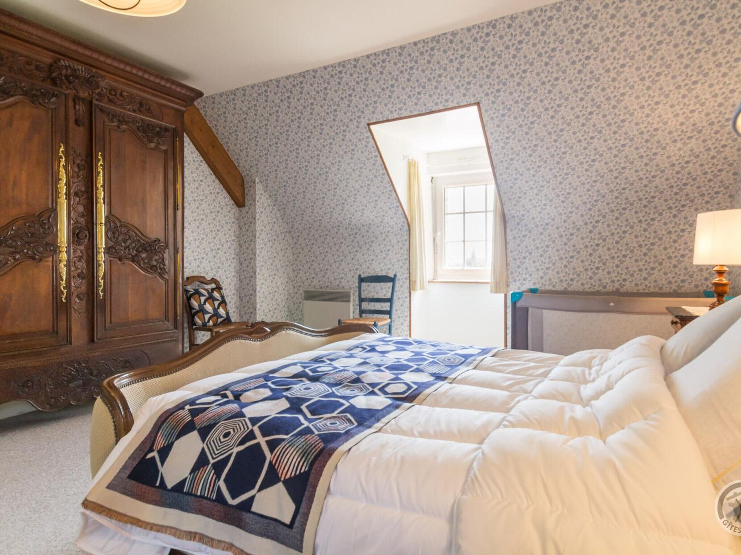 A bedroom with a bed, a wooden wardrobe, a chair, and a window.