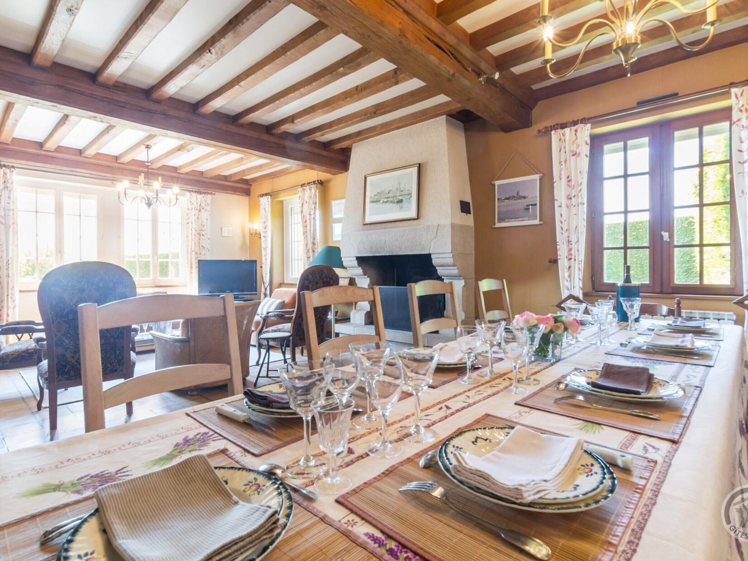 A dining area with a long table set for a meal, wooden beams on the ceiling, and large windows with curtains.