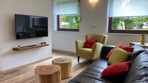 A modern living room with a flat-screen TV mounted on the wall, a wooden shelf below, a yellow armchair with red cushions, a black leather sofa with red and black cushions, and two wooden stools. - Modern vacation home that leaves nothing to be desired at the Eifel National Park (Hürtgenwald)