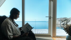 A person reading a book by a large window with a view of the sea and rocky landscape.