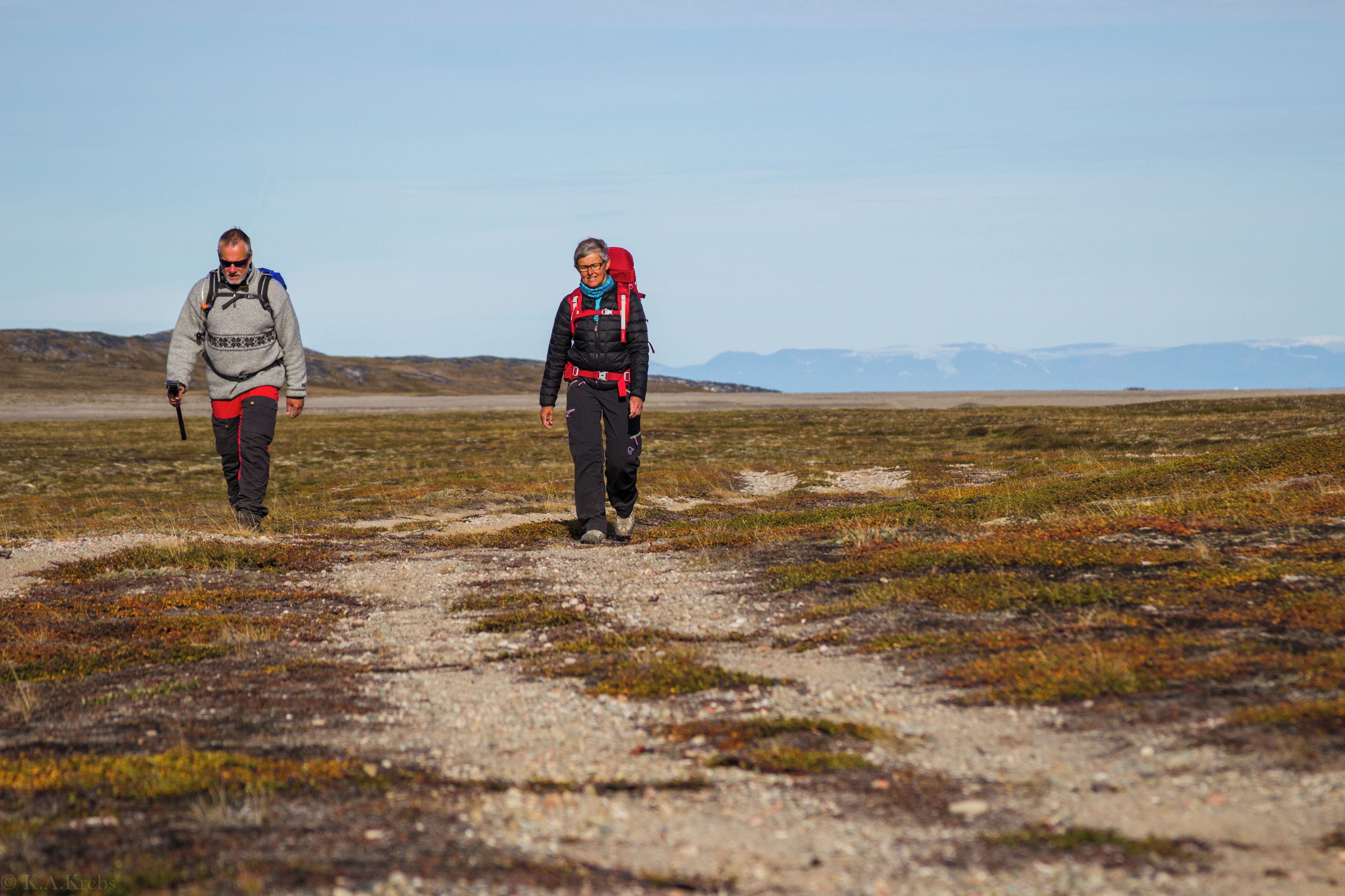 Two people walking on a gravel path in a flat, open landscape with distant mountains.