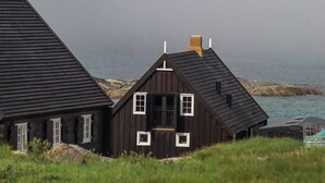 A dark wooden house with a steep roof, white window frames, and a chimney. It is situated near a rocky coastline with the sea in the background.