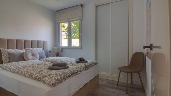 A bedroom with a large bed, a bedside table, a chair, a window with blinds, and a door.