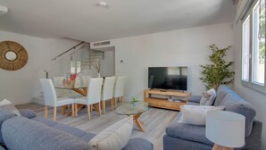 A modern living room with a glass dining table, white chairs, a grey sofa, a TV, and a staircase leading to an upper floor. - Spacious house just steps from the beach 2 min walk (Los Alcázares)