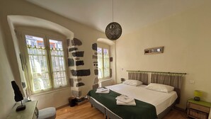 A bedroom with a large bed, a desk, and a chair. There are two windows with curtains, a stone wall, and a hanging light fixture.