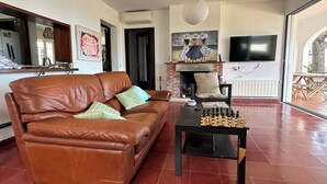 Een woonkamer met een bruine leren bank, een schaaktafel, een open haard, een televisie en een groot raam.